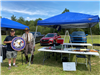 Vendor Set Up At Their Table Holding a Circular Sign with an Eagle in the Center at Copley Community Park on Saturday For Heritage Days 2025