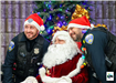 An officer Stands on either side of a sitting santa claus, between 2 Christmas trees