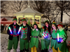 A group of high school aged kids are dressed in elf costumes holding light up wands, in the background is snow on the ground and a white gazebo
