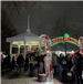 A group of people line up outside on the snow covered ground, the line goes under an illuminated holiday sign saying "Happy Holidays" the line goes towards a white gazebo where santa claus is taking pictures with children