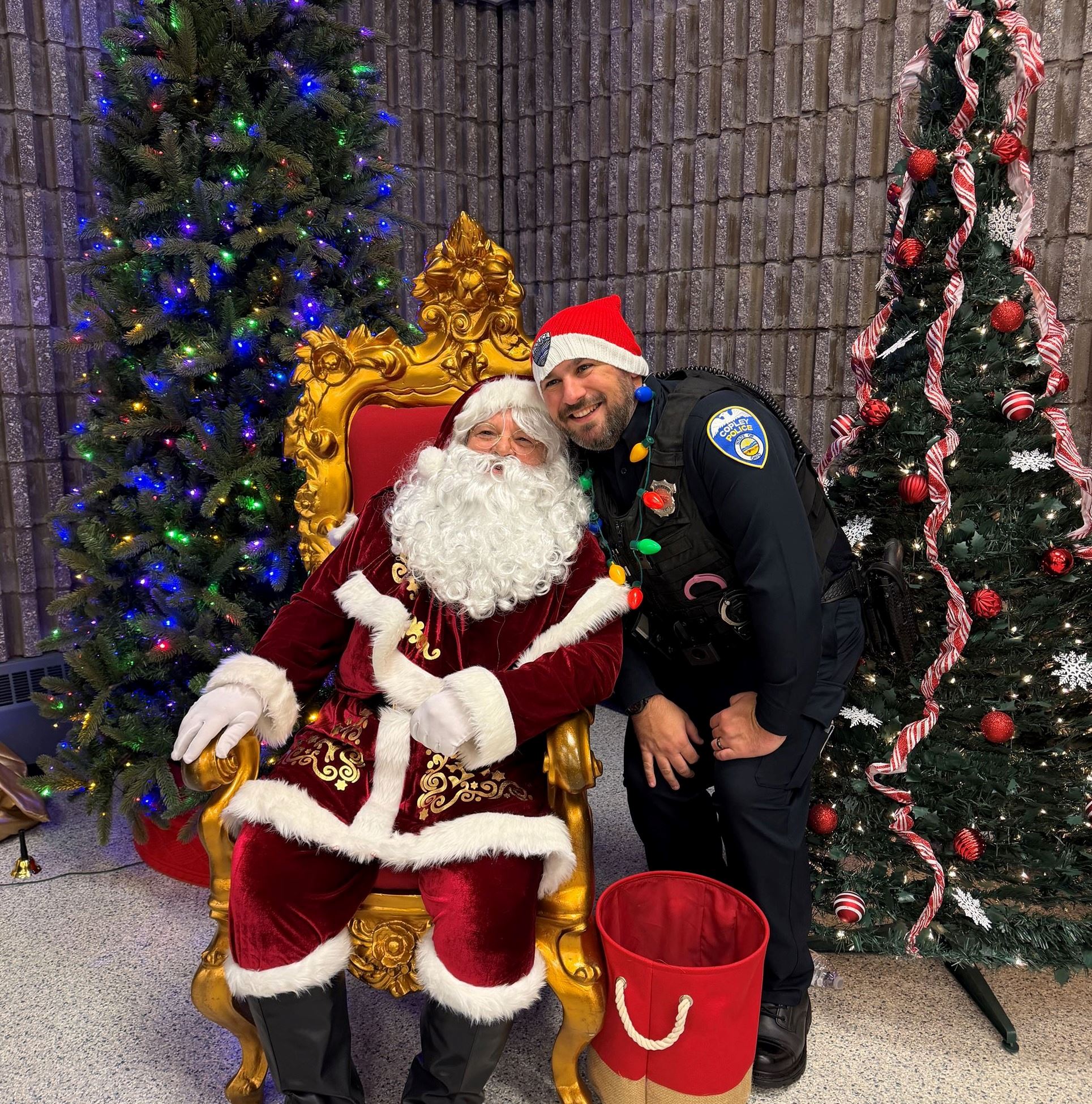 An officer stands next to a sitting Santa Claus, between 2 Christmas Trees