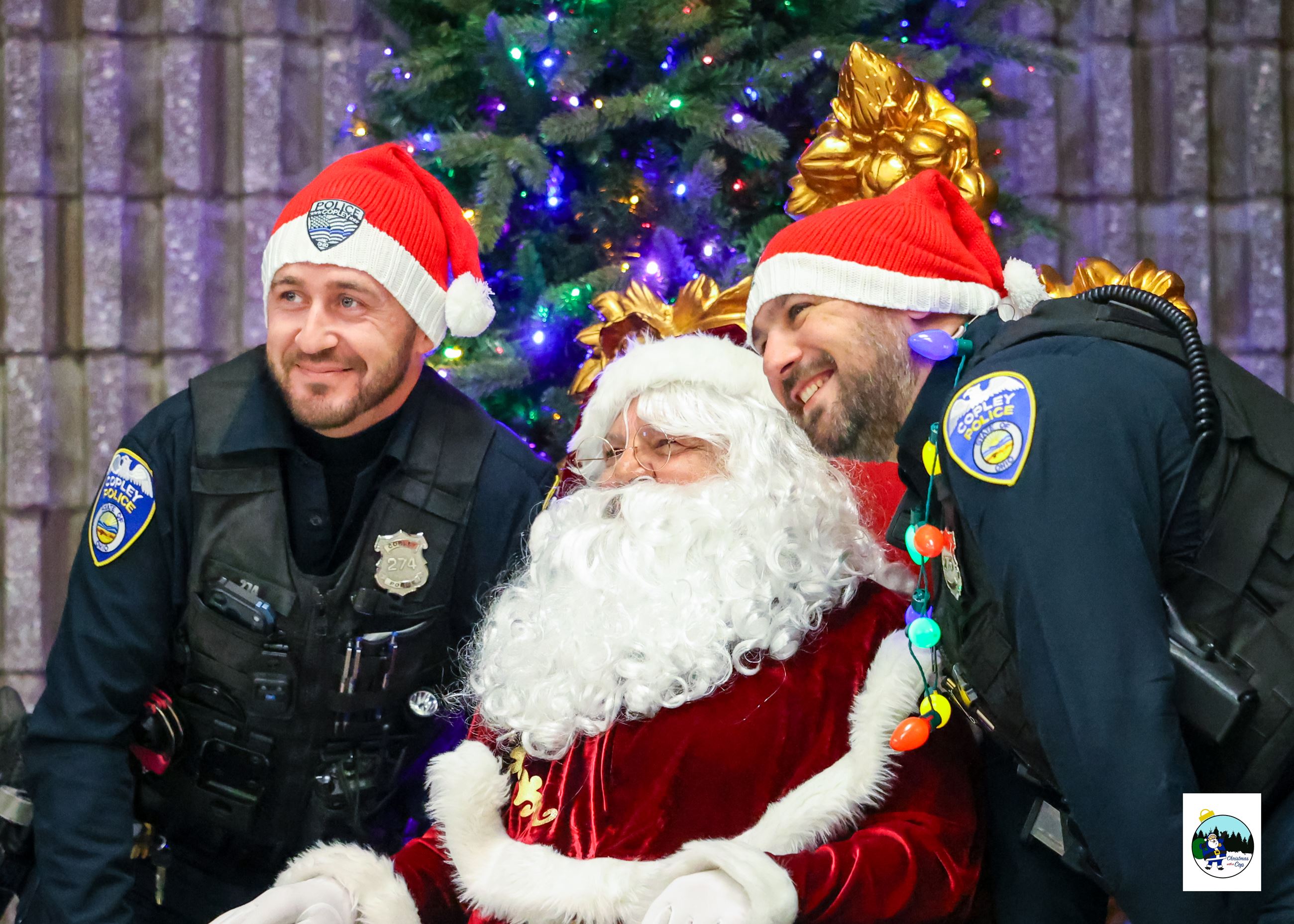An officer Stands on either side of a sitting santa claus, between 2 Christmas trees