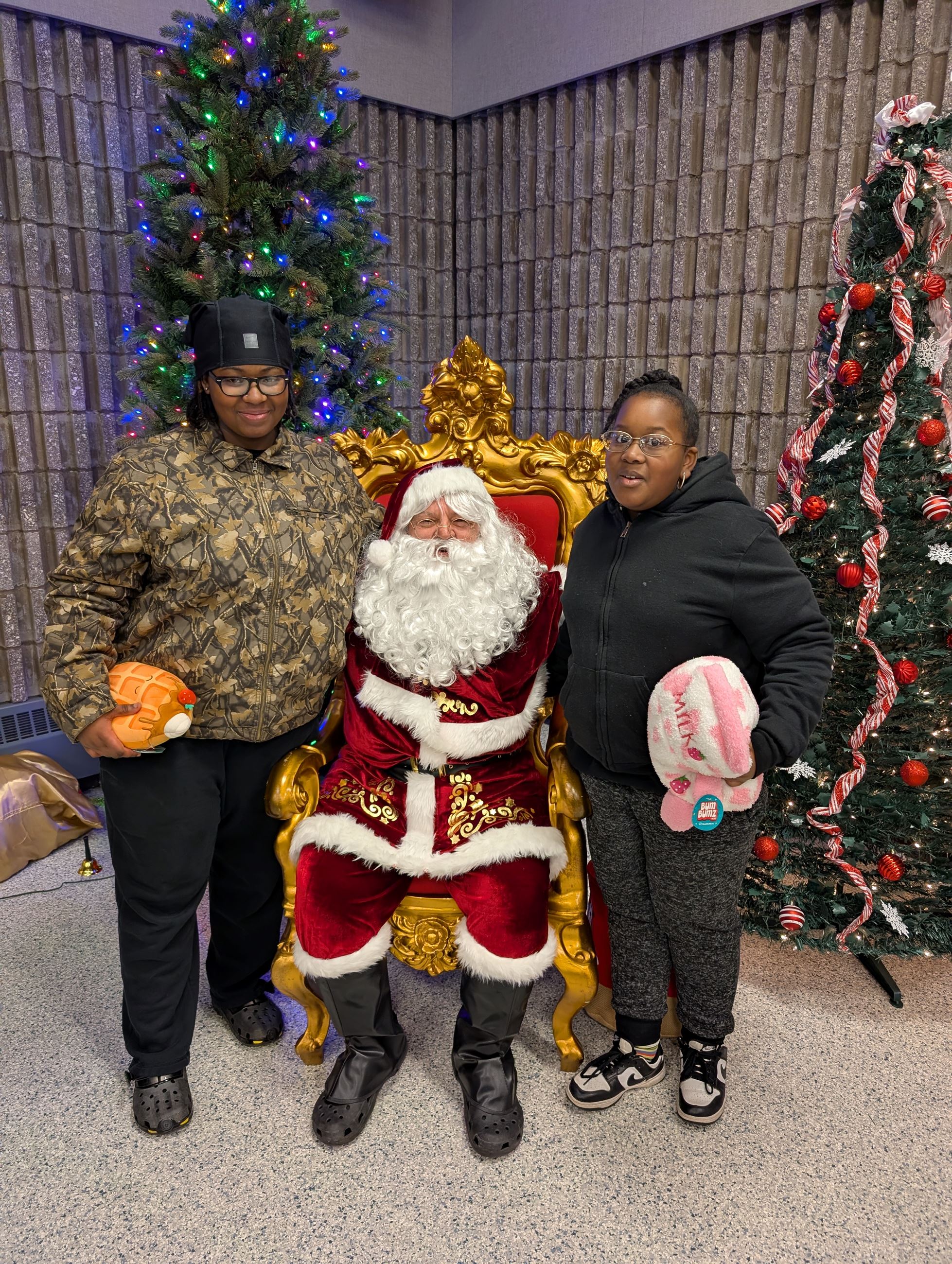 A child stands on either side of Santa Claus, between 2 christmas trees