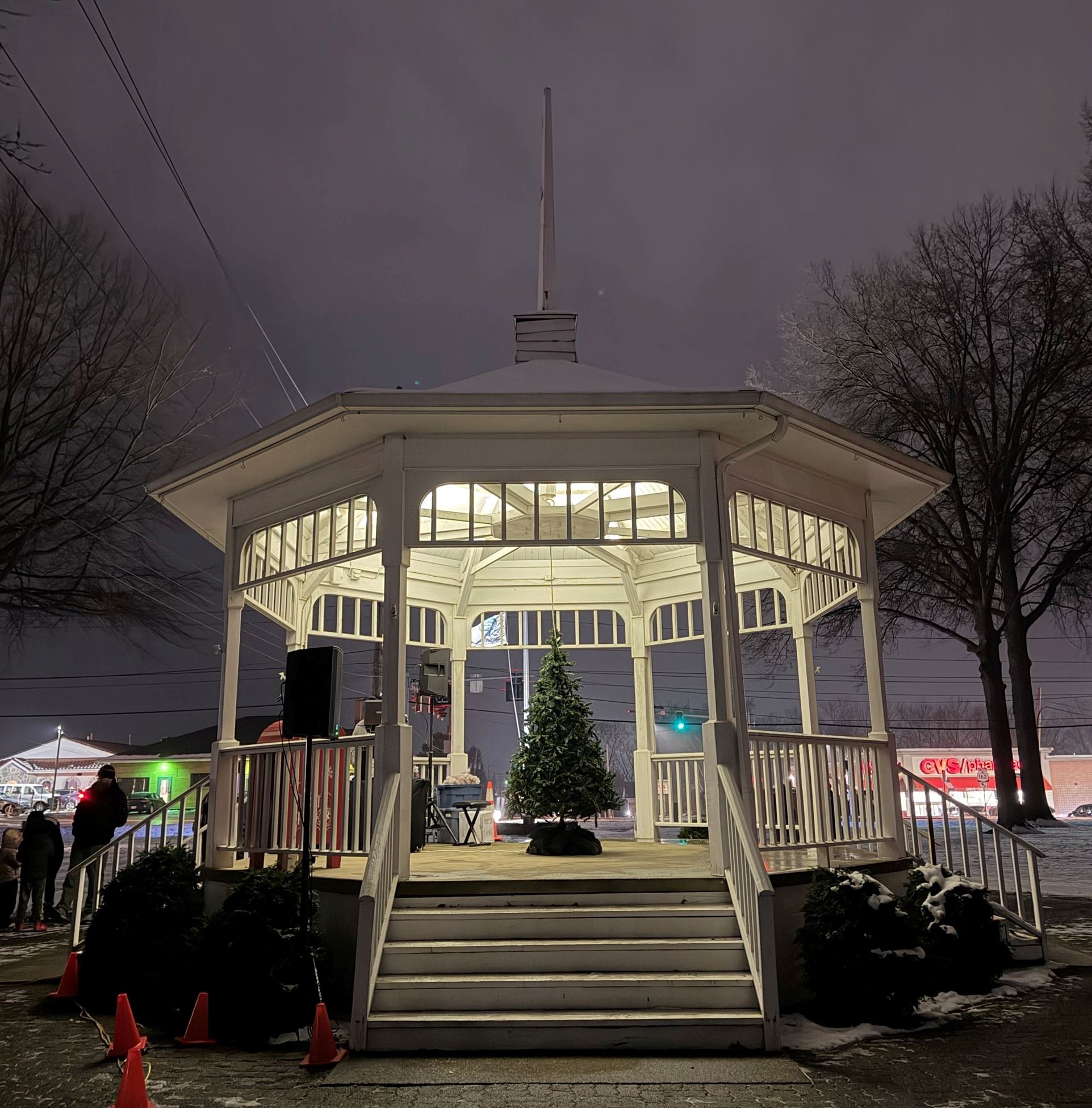 A white gazebo surrounded by snow on the ground, a christmas tree is in the center of the gazebo but is not illuminated