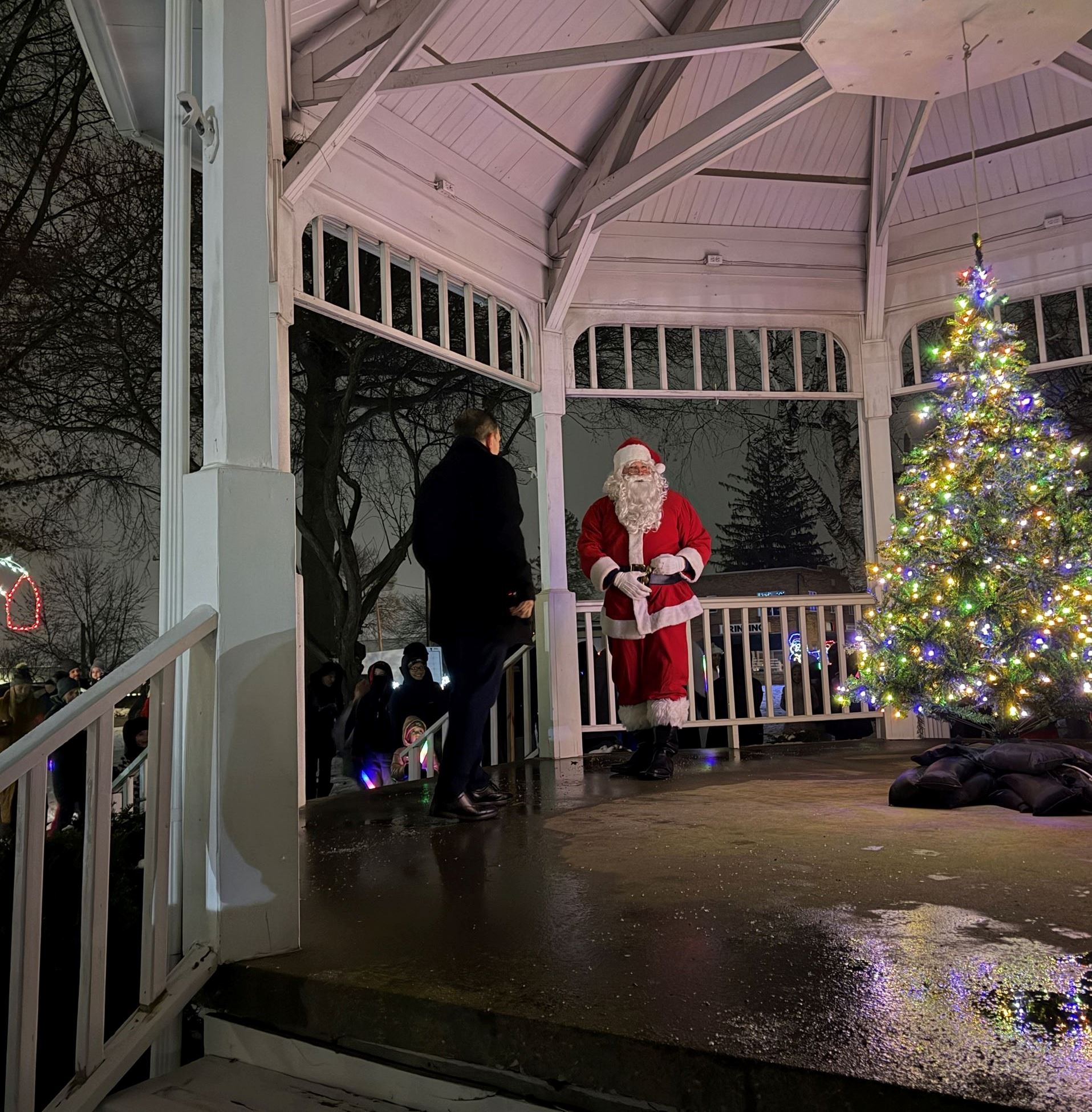 A man is seen standing on the Gazebo stage with Santa Claus and a Christmas Tree